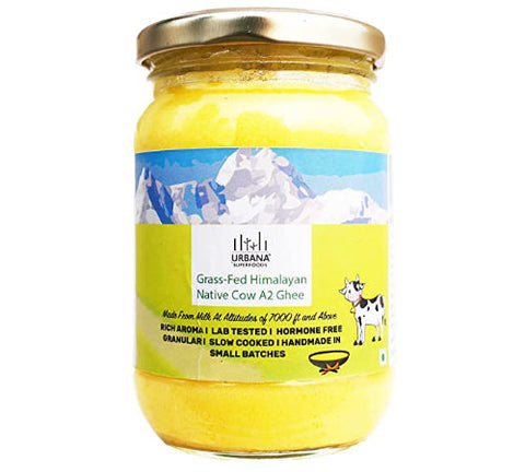 An image depicting a glass jar of Urbana Superfoods Grass-Fed Himalayan Native Cow A2 Ghee, filled with bright yellow granulated ghee, featuring mountain peaks and a cow illustration on the label, symbolising purity, Himalayan sourcing, and traditional handmade preparation.